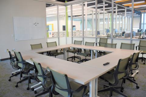Six tables arranged into a square with twelve chairs and a large mounted whiteboard. Large glass windows looking out into the library. 