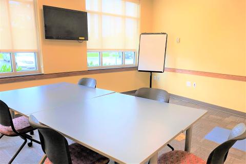 Shows a large room with a mounted television and a standing white board. There are two tables pushed together with five chairs surrounding the table.