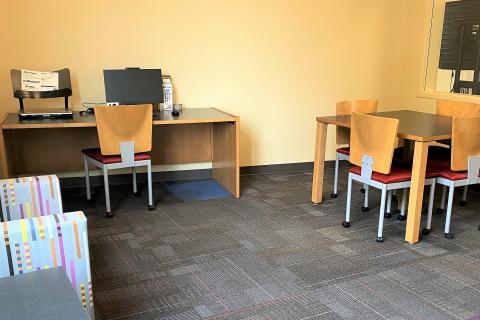 The study room has yellow walls with mostly blue geometic-print carpet. A desk with a rolling chair and desktop computer are located along the far wall, with a small table and four chairs located on the perpendicular wall to the right. Two armchairs and another small table are located on the wall opposite the table and chairs. 