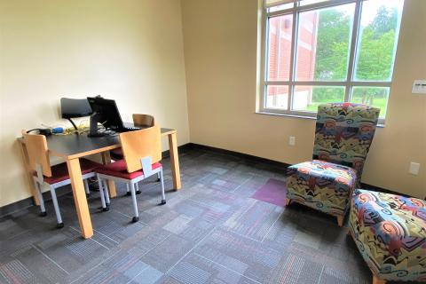 The study room has yellow walls with multi-colored geometic-print carpet. A small table with three chairs and a computer is pushed against one wall. Two padded chairs are located in a corner across the room.