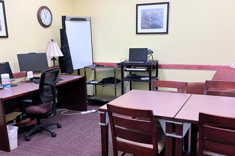 Shows partial view of the study room, which has yellow wall and maroon carpet. The room includes a desk area complete with a desktop computer and rolling chair plus a small table area that can seat upt to four comfortably. Additional equiptment, including an AV cart and whiteboard, are located in a corner. A wall clock is hung in the same corner.