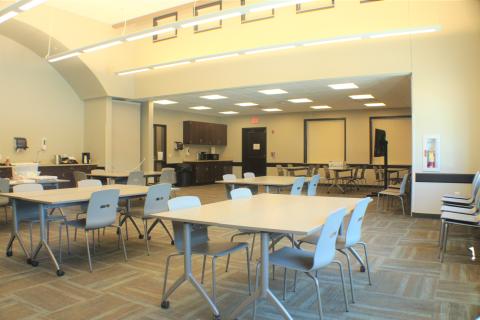 A large room with 12 tables and four chairs at each tables. Cabinets, sink, and microwave on back left corner, and a televeision on a rolling cart. 