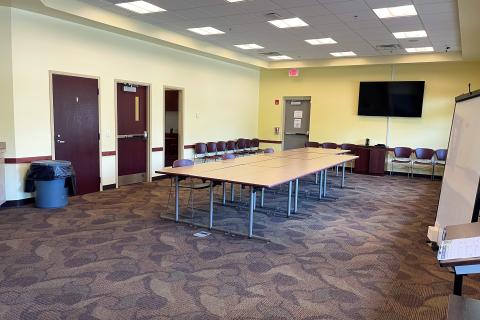 Image shows a room with yellow walls, patterned burgundy carpet, and dark wood. Long tables and chairs are gathered in the center of the space and a flat screen TV is mounted on the wall. In the right-hand corner, a white board and podium are visible.