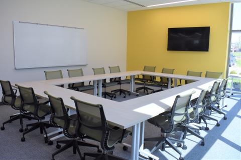 Shows a conference room with 8 tables and 16 chairs set up in an open square configuration. There is a large whiteboard mounted to the wall and a TV mounted to the wall.