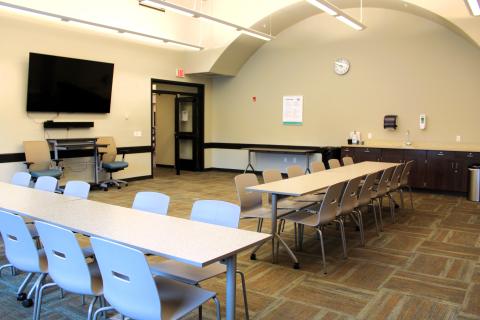 Images shows the front portion of Meeting Room 1 with rectangulat tables and chairs set up  to form and "L" facing a mounted TV. A sink and counter space are visible in the background.