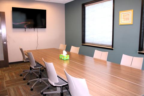 Image shows two sides of a beige room with a gray-blue accent wall and brown carpet. A large TV screen is mount on the narrower beige wall and in the center of the room, four rectanglular tables are pushed together to form a larger boardroom-style table. Eight taupe/beige plastic chairs are positioned at the table.