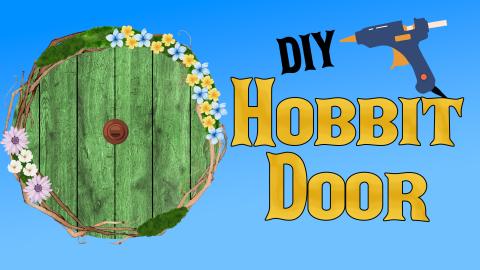 Image reads "DIY Hobbit Door" against a blue gradient background. A hot glue gun is above the title. To the left of the title is a green wooden circular door with a handle in the middle and a grapevine wreath with flowers surrounding the door.