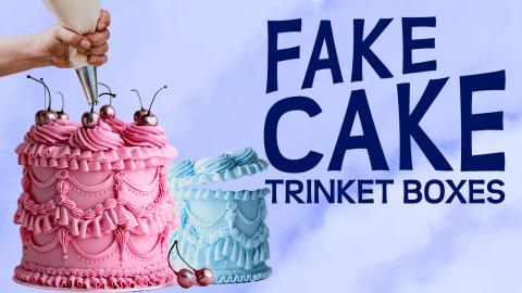 Image reads "Fake Cake Trinket Boxes" against a textured background. To the left of the title are two fake cake boxes and a hand holding a pipping bag. 