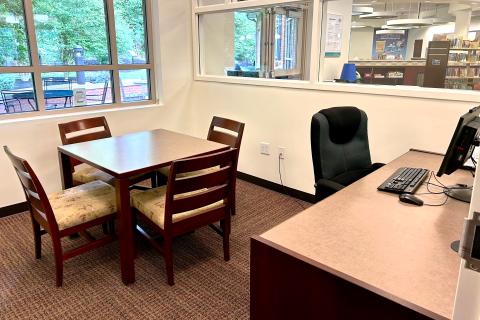 A large desk space with a rolling chair and desktop computer are located in the right corner of a room with beige walls and brownish-maroon carpet. A square table with four chairs also sits in view of a large window with a peaceful view of the back patio.