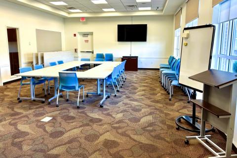 Image shows a room with off-white walls, patterned brown and burgundy carpet, and dark wood. Light wood tables and blue chairs are arranges in a larger rectangle in the center of the space and a flat screen TV is mounted on the wall. Additional tables and chairs are stores along the walls on either side of the configured tables. In the right-hand corner, a white board and podium are visible.