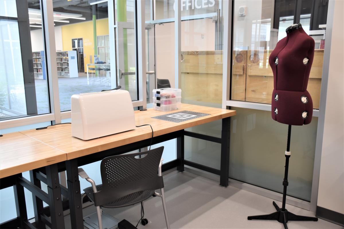 Image shows a sewing machine, a cutting mat on a desk, an empty mannequin, and a chair.