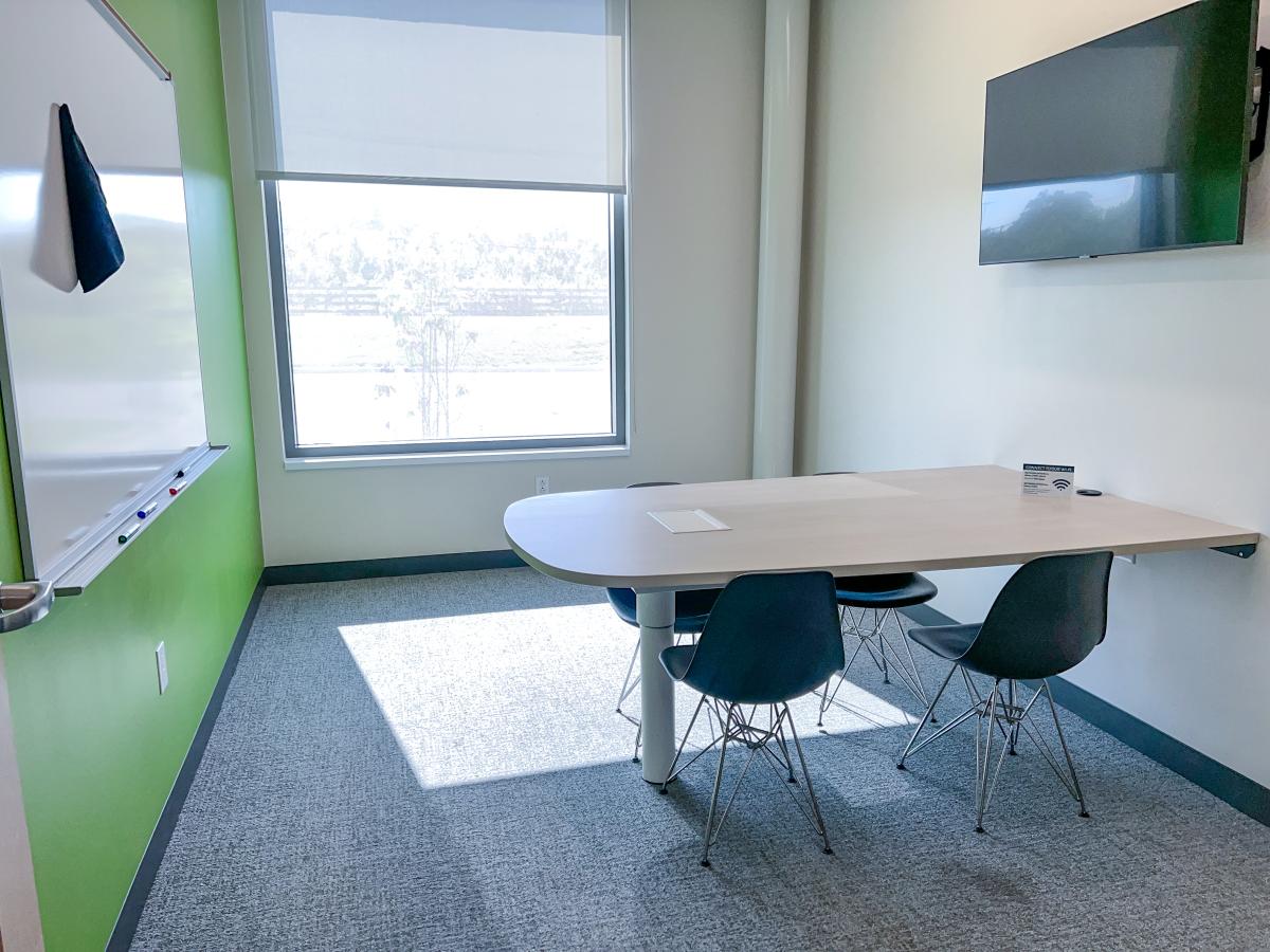 A table attached to the wall with four chairs with a mounted television and a mounted white board.